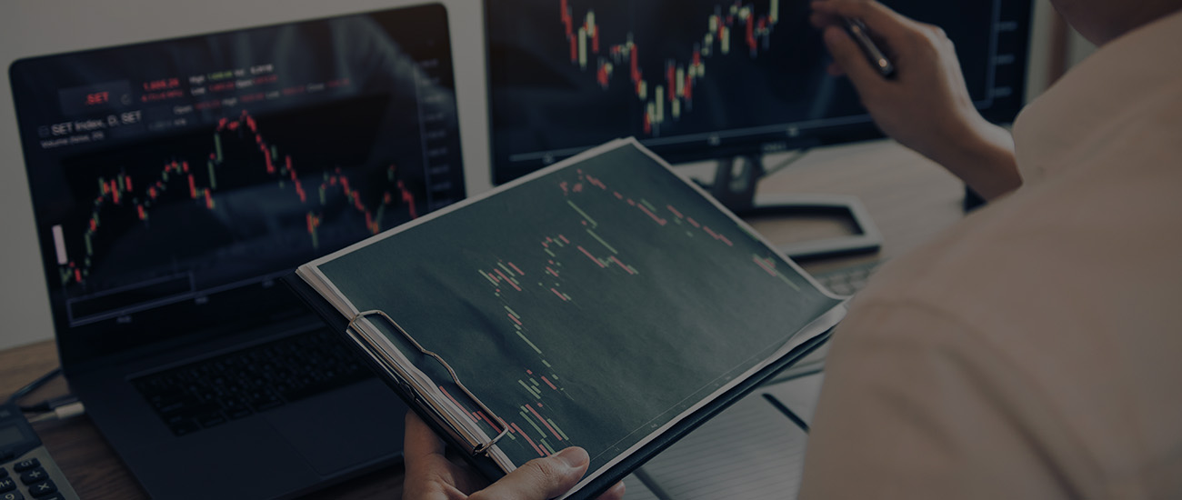 Close-up of stock market charts on computer monitors with a person holding a document showing additional financial data.